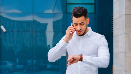 Portrait successful serious arabic hispanic business man spaniard boss leader wears white formal shirt stands outdoors speaks mobile phone cellphone answers call looks at wrist watch checks late time