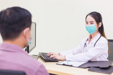 Professional Asian woman doctor records symptom of man patient in document at hospital, both of them wear medical face mask to protect Coronavirus (Covid 19)  while in examination room at hospital.