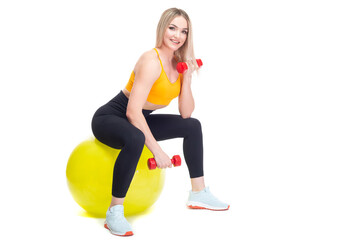 A beautiful, athletic, slim, smiling and cheerful woman in an orange top and black sweatpants performs exercises with dumbbells while sitting on a yellow fitball. Lifestyle concept with sports and gym