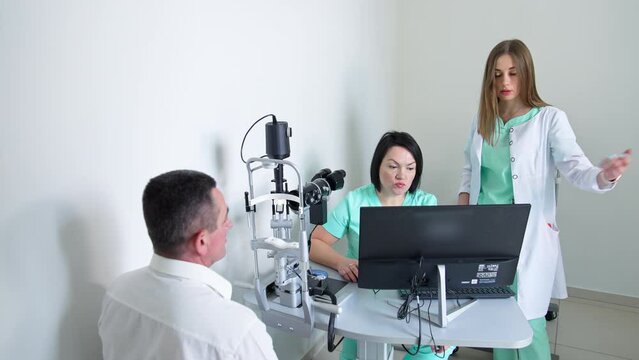Male patient sits in front of apparatus at oculist's cabinet. Two female doctors have discussion of case at computer's screen.