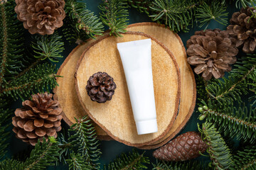 White plastic cosmetic tube between fir branches and pine cones on wooden piece top view. Mockup.
