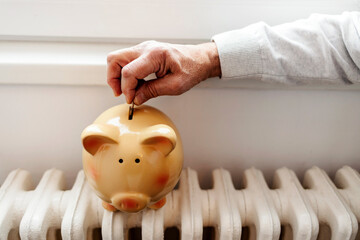 Energy saving concept. Men's hand inserts coin in piggy bank on a heating radiator. Saving money at the heating season time at home. Energy saving concept and efficiency house symbolic image.
