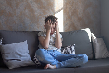 Obraz premium A teenage boy clutched his head with his hands, a teenage schoolboy is sitting in his room on the couch and is experiencing stress and resentment
