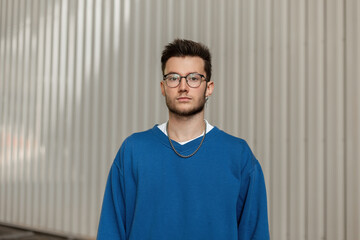 Fashionable young stylish hipster man with an earring and glasses in a trendy blue sweater on a...
