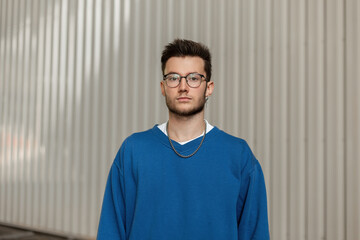 Fashionable young stylish hipster man with an earring and glasses in a trendy blue sweater on a gray background on the street