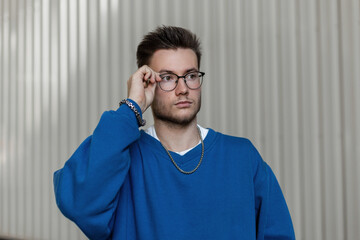 Stylish young hipster guy in a trendy blue sweatshirt wearing glasses on a metallic gray background...
