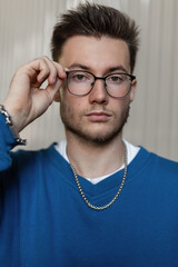 Obraz premium Stylish portrait of a handsome young hipster guy with hairstyle in a trendy blue sweater wearing glasses on the street