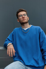 fashionable young male model hipster in a fashion blue sweatshirt with glasses stands outside a...