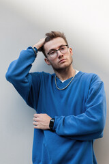 Fashion hipster man model with hairstyle and stylish eyewear in trendy blue sweatshirt stands and...