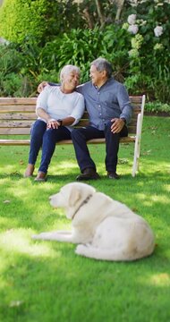 Vertical video of happy senior biracial couple with dog embracing in garden