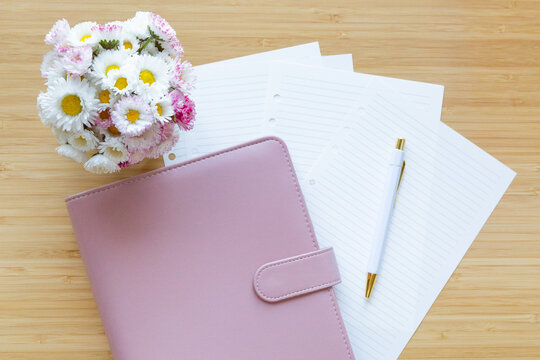 Flat lay, top view of a pastel pink planner, flower bouquet and stationery on a wooden desk.