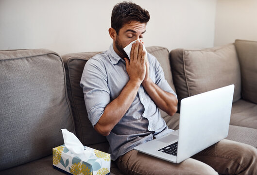 Hopefully he can mange to finish all his work today. Shot of a young businessman blowing his nose with a tissue while trying to work on his laptop at home.