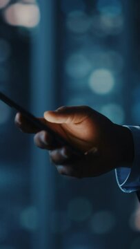 Vertical Screen. Night Office: Successful Black Businessman Wearing Suit Standing, Using Smartphone. Ambitious CEO Monitoring Growth of His e-Commerce Software. Focus on Hand with Mobile Phone