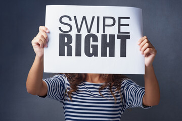 Its a match. Studio shot of a young woman holding a sign with swipe right printed on it against a gray background.