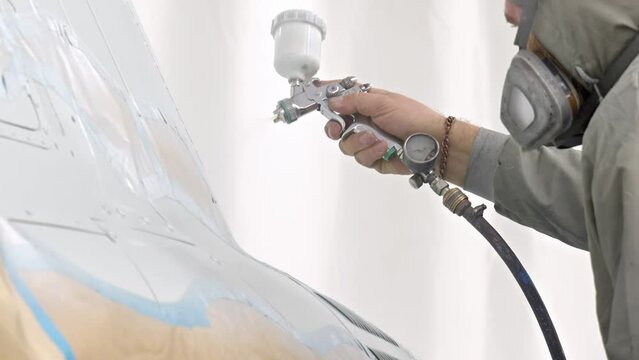Silver spray gun in spray booth in hands of painter. worker paints top of helicopter or car. White paint is sprayed close up in slow motion. worker restores aircraft.