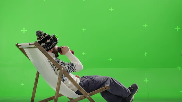 A man snowboarder siting in sun lounger and drinking hot drink. Guy in winter clothes enjoying mountain view. Imitation of ski resort on green screen in studio. Tracking points , chroma key 