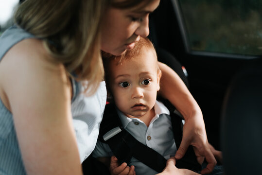 Young caucasian mother fastening her little son with safety belt in child seat inside car. Selective focus on child's face with frightened, excited expression