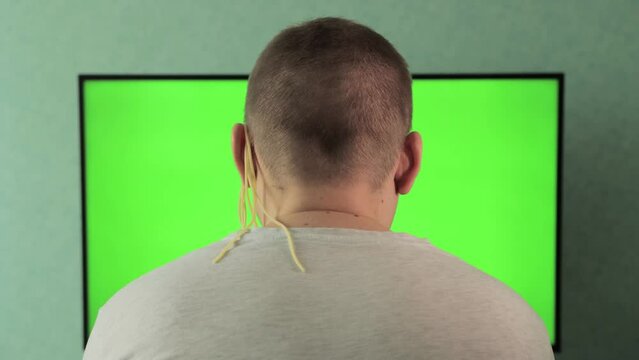A man in front of a chroma key green screen TV is watching TV. Removing noodles from the ears of viewers. The concept of deception and fecks on television. News channel