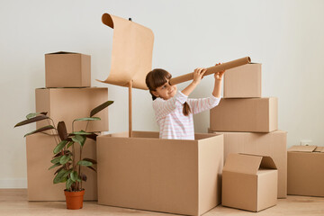 Charming dark haired little girl playing in handmade ship from cardboard box with sail, female child with spotting scope, posing surrounded with carton parcels.