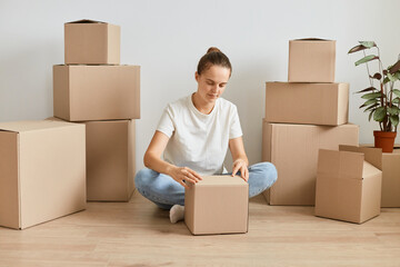 Indoor shot of attractive woman wearing white t shirt and jeans sitting on floor surrounded with cardboard boxes and unpacking carton parcel with her personal belongings.