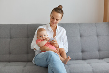 Portrait of attractive young adult woman wearing white shirt and jeans sitting on sofa with baby, mother feeding her daughter from the bottle, expressing positive emotions.