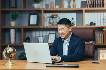 Portrait of a successful Asian businessman in a classic office working on a laptop, smiling - obrazy, fototapety, plakaty