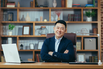 Obraz premium Portrait of a successful Asian businessman in a classic office working on a laptop, smiling