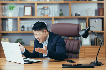 Frustrated and angry asian businessman working on laptop in office, boss shouting at laptop screen