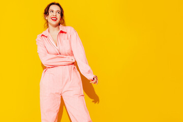 Fototapeta na wymiar Young beautiful smiling female in trendy summer pink overalls. Sexy carefree woman posing near yellow wall in studio. Positive model having fun and going crazy. Cheerful and happy