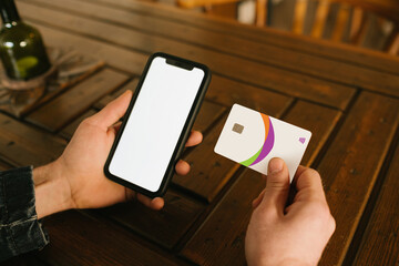 Young man sitting on a cafe with credit card and phone in hands doing purchases during online shopping. Online payment. Man holding smartphone with blank screen and credit card, making transaction