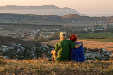 Fototapeta na wymiar An elderly couple sits on a mountain with their backs with a beautiful view of the mountains and the sea in the distance.