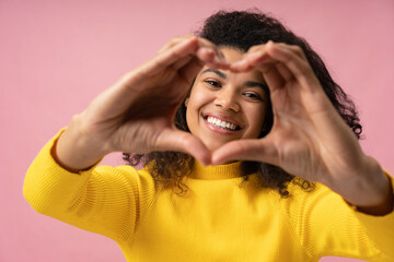 Beautiful African American woman with toothy smile showing heart shape near face isolated on pink background. Health care, dental concept   