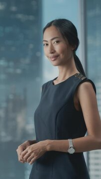 Vertical Portrait of an Asian Businesswoman in Stylish Black Dress Using Laptop Computer, Posing Next to Window in City Office. Confident Female CEO Smiling. Successful Diverse Business Manager.