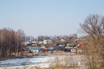 A small frozen river, rural houses in the background. Spring, snow melts, puddles and dry grass all around. Day, cloudy weather, soft warm light.
