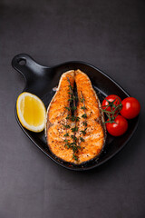 Grilled salmon with cherry tomatoes, thyme and lemon on a small black grill pan. Fried fish is a traditional dish of home cooking and restaurants. Black background, selective focus, close-up.