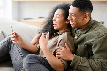 Side view of couple in love are sitting on the couch at home and using smartphone for talking online, video meeting with a friends