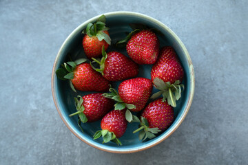 Red fresh strawberries with green leaves in plate on gray background. Top view