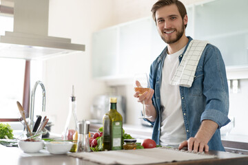 Handsome young man in casual wear drinking wine while standing in the kitchen at home.
