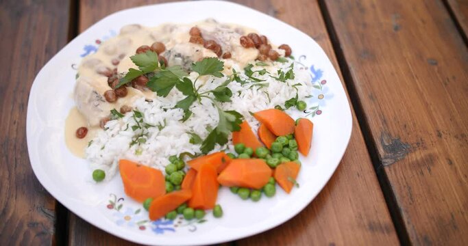 Top view of rice with white sauce and raisins along with boiled meat with carrots and peas on a white plate over the wooden table. cilantro on the top.