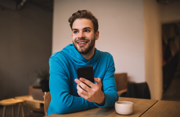 Happy Caucasian male with cellular technology enjoying positive break pastime in cafe interior, cheerful millennial man holding digital smartphone gadget and smiling during leisure in coffee shop