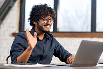 Cheerful attractive Indian guy using laptop for video communication. Guy in glasses sitting at the...