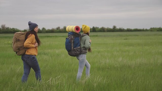 happy girls walk across green field with backpacks. two female friends resting together journey. weekend nature with travel bags. female adventure vacation. teenagers go to the mountains on the grass.