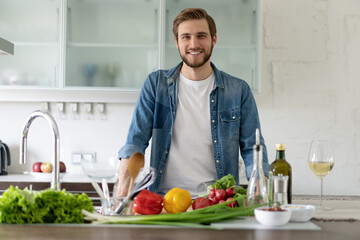 Handsome smiling young man leaning on kitchen counter with vegetables and looking at camera.