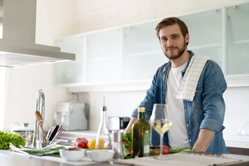 Fototapeta na wymiar Handsome smiling young man leaning on kitchen counter with vegetables and looking at camera.