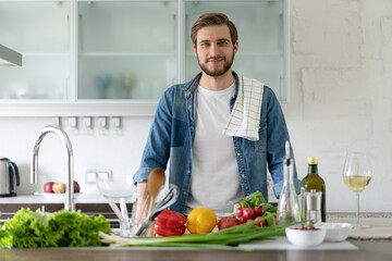 Handsome smiling young man leaning on kitchen counter with vegetables and looking at camera.