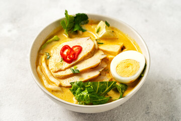 Asian laksa soup with chicken meat in a ceramic bowl on a culinary background. Spicy soup with coconut milk broth, rice noodles and egg in a plate on the kitchen table	