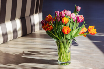 A bouquet of yellow and pink tulips in a glass vase stands on the floor in bright sunlight with contrasting shadows
