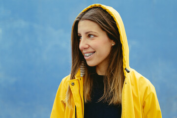 Close-up side portrait of blonde woman wearing a raincoat. Horizontal view of caucasian woman in the street with yellow raincoat isolated on blue background. People lifestyles.