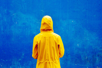 Rear view of woman wearing a yellow raincoat isolated on blue background. Horizontal mid waist view of unrecognizable person wearing raincoat under the rain on blue background. Seasonal backgrounds.