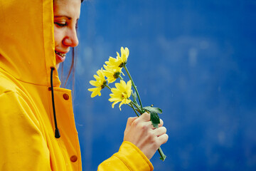 Side view of woman wearing a yellow raincoat smelling flowers. Close-up view of woman under the rain holding a yellow bouquet of daisies flowers isolated on blue background.